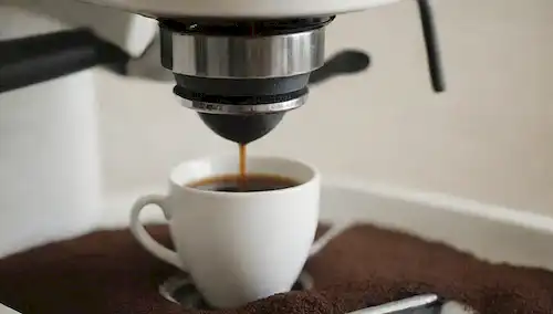 Una toma de cerca, fondo blanco, máquina de café limpia y vieja, lente de cámara visible, cesta de filtro abierto, café marrones derramado, depósito de agua parcialmente lleno, agujeros de vapor ligeramente sucio