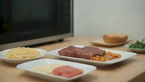 Un plato con comida congelada, un horno de microondas en el fondo, un recipiente vacío, una cuchara, un vaso de agua, una tabla de cortar y un cuchillo