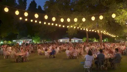 Una cálida noche de verano, un parque histórico en Madrid, un ambiente animado, gente reunida, linternas coloridas, luces parpadeantes, árboles verdes, flores vibrantes, terrazas con cómodos asientos, modernas y elegantes decoraciones, una banda que interpreta música en vivo, las ondas sonoras visualizadas como ondas onduladas