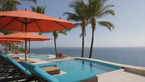exterior hotel de 5 estrellas, sillas de playa con el sol, piscina infinita, palmeras, aguas azules del océano, asientos confortables de salón, barandillas de cristal, arquitectura moderna, sombrillas de colores vibrantes, ambiente sereno, impresionante ciudad skyline