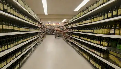 un pasillo de cocina con hileras de botellas de aceite de oliva, una variedad de marcas y tamaños en estantes, un carrito de compras o canasta en primer plano, un fondo de supermercado brillante y moderno