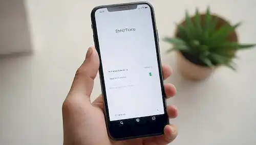 Un iPhone renovado en un fondo blanco con un icono de marca de verificación sobre él y un icono de sobre debajo de él; un efecto gradiente sutil para separar el objeto del resto; una mano pequeña que sostiene el teléfono, transportando un sentido de propiedad o manipulación; enfoque suave para evitar bordes agudos; tono de color cálido para sugerir comodidad; ligera borda alrededor de los bordes para crear profundidad