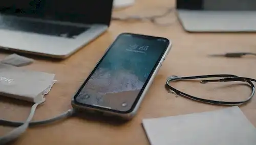 3D-rendered iPhone con casquillo usado, rasguños en la pantalla, logotipo ligeramente decolorado, espalda agrietada y pliegues débiles en los lados, sentado encima de un escritorio desordenado con papeles dispersos, cables y dispositivos en el fondo, todo en colores mudos