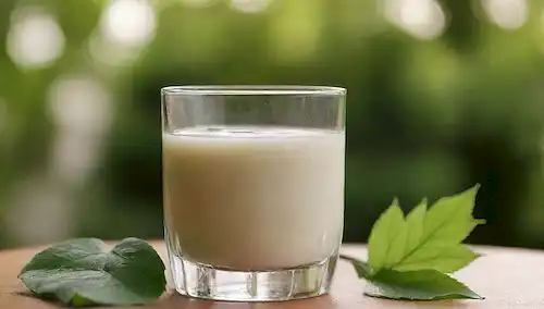 un refrescante vaso lleno de líquido blanco cremoso, rodeado de hojas verdes vibrantes, situado en un fondo soleado cálido, con unos pocos cubos de hielo y una rociada de canela en la parte superior
