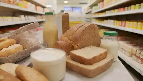 Un primer plano de un carrito lleno de diversos productos alimenticios, incluyendo productos sin lácteos y sin lactosa, como alternativas de queso vegano, botellas de leche de almendra, panes sin gluten y una cesta de fruta fresca En el fondo, una imagen borrosa de una estantería o pasillo para proporcionar contexto