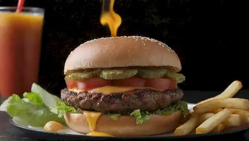 Un jugoso patito en una parrilla, rodeado de hojas crujientes de lechuga, rebanadas de tomate maduro, queso cheddar fundido, y un bollo dorado, con un lado de patatas fritas crujientes y un refrescante vaso de soda