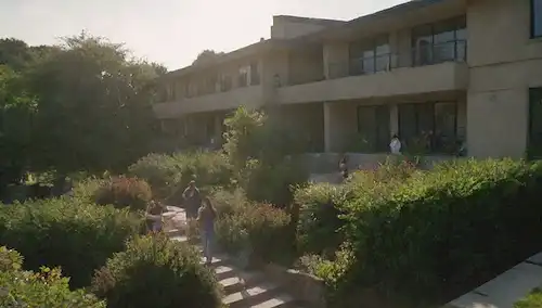 complejo de apartamentos con zona común al aire libre, trabajadores caminando escaleras, empleados trabajando en el techo, equipos de mantenimiento dispersos, jardín comunitario en el fondo, sol filtrando a través de árboles, exuberante vegetación rodeando edificio