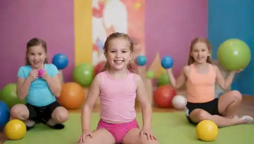 3-6 niños de 4 a 12 años, sonriendo y riendo, sosteniendo pequeñas campanas, timbres o bandas de resistencia, sentado o de pie en un fondo colorido con bolas de ejercicio, vigas de equilibrio o bloques de espuma