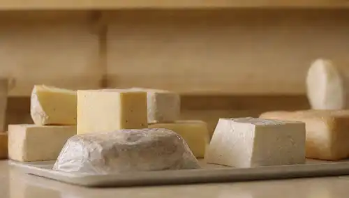 Una foto de un estante apilada con varios tipos de queso, algunos envueltos en envoltura plástica, otros en recipientes herméticos, en un fondo cálido con una encimera de cocina borrosa, iluminada por luz natural suave, con unas cuantas galletas o rodajas de pan colocadas al lado
