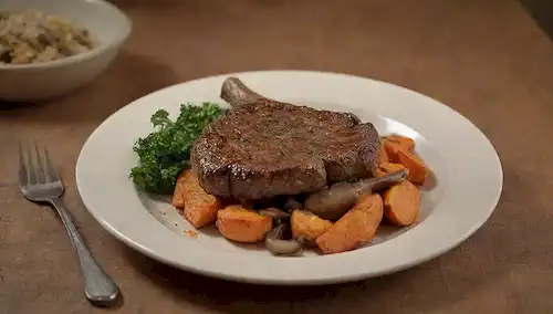 un plato con una cuna de cordero cocido, rodeado de hongos saltados, un lado de la batata, una rociada de perejil fresco, en una mesa de madera rústica con un fondo vintage, iluminación caliente, y algunas migajas dispersas alrededor