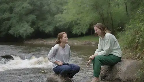 Un par de pantalones hechos de fibras naturales siendo lavados en un río con una pequeña cantidad de burbujas detergentes, un pulverizador que libera plaguicida cerca, una persona sonriente con ropa de fibra sintética sentada sobre una roca en el fondo, plantas verdes y árboles creciendo alrededor de la orilla del río, un contraste entre flujos de agua limpia y contaminada, un sutil indicio de contaminación en el aire