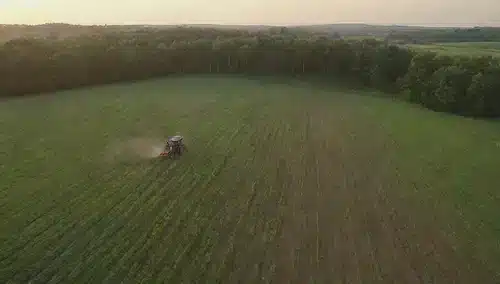 Un paisaje verde con una mezcla de cultivos y equipos agrícolas, agricultores inspeccionando campos con tabletas, drones volando sobre la cabeza, sensores unidos a plantas, tractores equipados con tecnología de precisión, filas de árboles con rociadores GPS habilitados, y un fondo de puesta del sol o amanecer con nubes