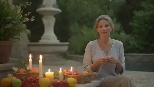 Una mujer serena en su vida media sentada en un entorno natural, con una canasta llena de frutas frescas y verdes frondosos a su lado, rodeada de elementos calmantes como velas, flores o una fuente de agua, mostrando un estilo de vida equilibrado