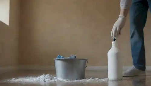 Una persona que lleva guantes y una máscara que sostiene una botella de spray con una piedra en el fondo, un cubo con polvo blanco, un cepillo de escob y un paño de microfibra, rodeado de paredes de color neutro con algunas piedras decorativas